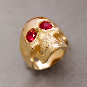 May include: A gold skull ring with two red ruby eyes. The ring is detailed with a skull face and teeth.