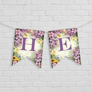 Op de afbeelding: Twee wimpelbanners met de tekst "HE" hangen tegen een witte bakstenen muur. Elke banner heeft een grote paarse letter, omgeven door een bloemenontwerp met paarse seringen, gele narcissen en zonnebloemen.