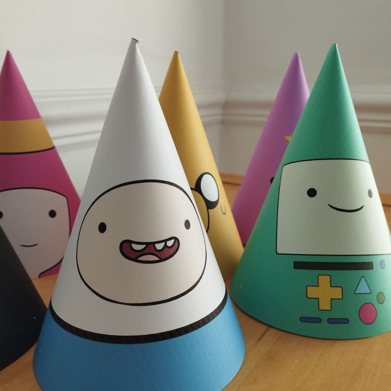 Może przedstawiać: Kolekcja kolorowych czapek na przyjęcia z postaciami z kresk&oacute;wek. Czapki mają kształt stożka i przedstawiają postacie takie jak Finn, Jake, BMO i Princess Bubblegum. Czapki są wykonane z papieru i mają niebieską, białą, ż&oacute;łtą, r&oacute;żową i zieloną kolorystykę.