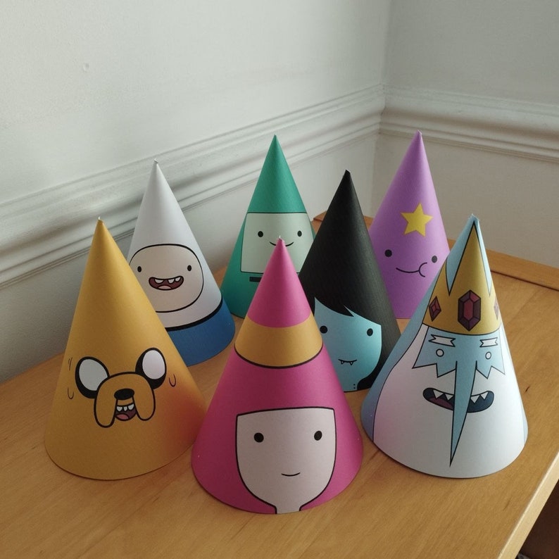 Może przedstawiać: Kolekcja siedmiu kolorowych czapek imprezowych z postaciami z serialu animowanego Adventure Time. Czapki są w kształcie stożka i mają nadrukowane twarze postaci, w tym Finna i Jake'a. Ułożone są na drewnianej powierzchni.