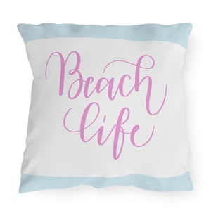 May include: A white throw pillow with a light blue stripe at the top and bottom. The pillow has pink cursive text that says "Beach life".