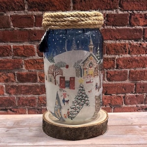 Könnte beinhalten: Ein dekoratives Glas mit einer Winterlandschaftsillustration. Das Glas zeigt ein verschneites Dorfdesign mit Kirche, Bäumen und Menschen. Es hat ein Seildetail am oberen Rand und steht auf einem Holzsockel.