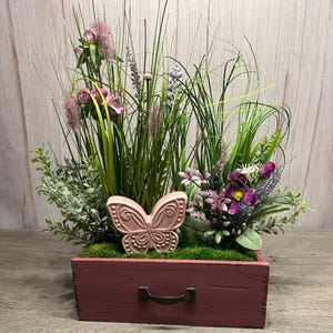 Könnte beinhalten: Eine rosafarbene Keramik-Schmetterlingsfigur sitzt in einem rosafarbenen Holzkasten, der mit grünem Gras und rosa Blumen gefüllt ist.