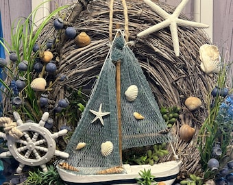 May include: A nautical-themed wreath featuring a small sailboat with a blue net sail, seashells, and a white ship's wheel. The wreath is made of woven branches, adorned with faux blueberries, greenery, and a starfish, creating a coastal aesthetic.