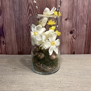 Könnte beinhalten: Eine transparente Glasvase gefüllt mit weißen und gelben Blumen, grünem Moos, kleinen gesprenkelten Eiern und einer Schnecke. Die Vase steht vor einem Holz Hintergrund, wodurch eine natürliche und dekorative Anordnung entsteht.