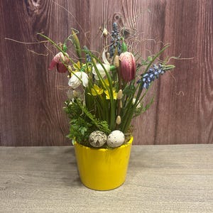 Könnte beinhalten: Ein leuchtend gelber Keramiktopf gefüllt mit einer Auswahl an künstlichen Blumen, darunter Tulpen, Narzissen und Blaubeeren. Zwei gesprenkelte Eier liegen auf dem Rand. Die Anordnung befindet sich vor einem hölzernen Hintergrund und schafft eine Frühlingsausstellung.