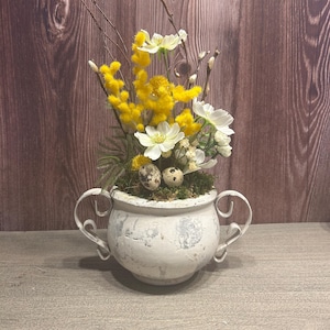 Könnte beinhalten: Ein weißer Keramiktopf mit Used-Look und zwei Griffen, gefüllt mit gelben und weißen Blumen, Grünzeug und zwei gesprenkelten Eiern. Die Anordnung steht auf einer grauen Oberfläche mit einem Holzgrund.