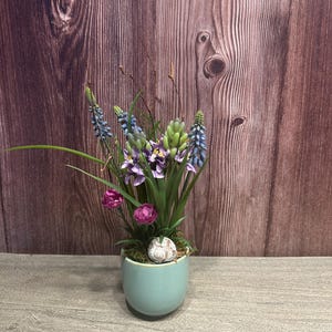 Könnte beinhalten: Ein kleiner, hellblauer Keramiktopf gefüllt mit künstlichen Blumen. Das Arrangement umfasst lila, rosa und blaue Blüten, mit grünem Laub und einem kleinen dekorativen Stein. Der Hintergrund ist eine Holzwand.