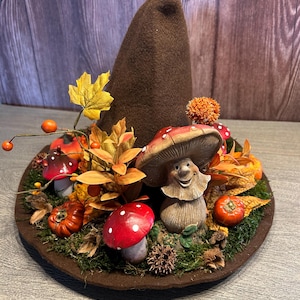 Könnte beinhalten: Ein brauner Hexenhut aus Filz, verziert mit herbstlichen Elementen. Der Hut hat rot-weiße Pilzfiguren, orangefarbene Kürbisse und Herbstlaub. Der Hut ist auf einem Bett aus grünem Moos platziert.