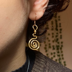 Peut inclure: Boucle d'oreille en fil d'or avec un motif en spirale. La boucle d'oreille est de style pendant, avec un crochet en haut et une spirale tourbillonnante en bas. La boucle d'oreille est portée à l'oreille.