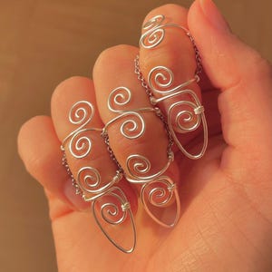 Peut inclure: Anneaux en fil d'argent au design unique. Chaque anneau présente une série de volutes et est relié par une fine chaîne. Les anneaux sont conçus pour être portés aux doigts, ajoutant une touche d'originalité.