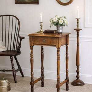 May include: A wooden side table with a single drawer, adorned with a brass pull. The table is flanked by a tall candlestick and a shorter one, both holding lit candles. A vase of white flowers and a book sit atop the table.