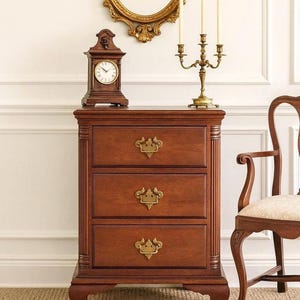 May include: A three-drawer wooden chest with brass hardware. A small wooden clock and a brass candelabra with three candles sit on top. A gold-framed mirror hangs on the wall behind the chest. A wooden chair is to the right.