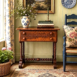 May include: A wooden side table with two drawers featuring round knobs and bobbin-turned legs. The table holds a ceramic vase with blue floral patterns, a brass candlestick, and two stacked books titled A Year in the English Countryside.