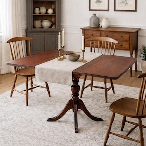 May include: A wooden dining table with two drop leaves extended, surrounded by four wooden chairs. The table is set with a cream-colored runner, candles, and a bowl. The room has a neutral color palette with a rug and a cabinet.