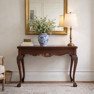 May include: A dark brown console table with ornate carved legs and a decorative floral accent. A blue and white vase with flowers, a lamp with a cream shade, and a gold-framed mirror are on the table. A small box and books are also on the table.