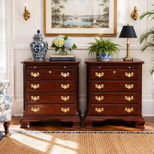 May include: Two matching dark wood chests of drawers, each featuring four drawers with ornate brass pulls and a keyhole. The chests are topped with decorative items including a blue and white porcelain jar, a glass vase with hydrangeas, and a lamp.