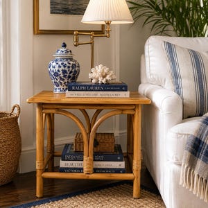 May include: A square bamboo side table with a lower shelf, featuring a brass swing-arm lamp, a blue and white porcelain ginger jar, a white coral piece, and several stacked books including titles by Ralph Lauren, India Hicks, and Mark D. Sikes.