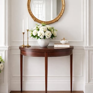 May include: A half-moon wooden console table with a dark finish, topped with a white vase of flowers, candles, and a decorative box. A gold-framed mirror hangs above. The room has white walls and trim.
