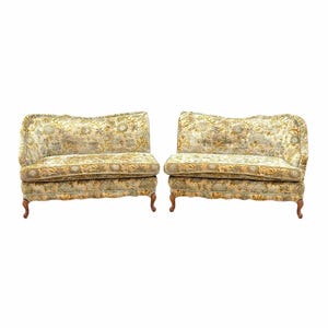 May include: A pair of loveseats featuring a floral pattern in shades of gold, beige, and muted green. Each piece has a curved backrest with ruffled trim, a single seat cushion, and four carved wooden legs with a curved, cabriole-style design.
