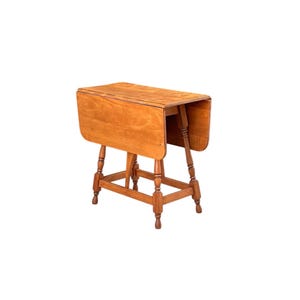 May include: A light brown, wooden drop-leaf table. The table has a rectangular top with two drop leaves, supported by turned legs and a connecting stretcher. The table is approximately 30 inches long when fully extended.