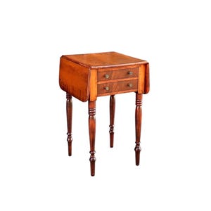 May include: A small, antique wooden side table with two drawers and a drop-leaf design. The table is made of dark brown wood and has four slender, turned legs. The drawers have small, round brass knobs. The table is a classic piece of furniture.