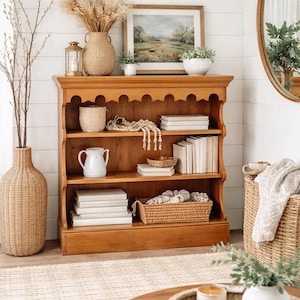 May include: A wooden bookshelf with three shelves, displaying books, decorative items, and a framed landscape painting. A large woven vase and a round mirror are also visible, creating a cozy, rustic interior design.