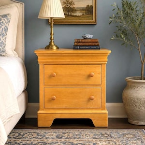 May include: A light-toned wooden nightstand featuring two stacked drawers with simple round wooden knobs. The piece has a rectangular top, a flat base with a cutout arch, and a smooth finish. It stands as a functional storage unit.