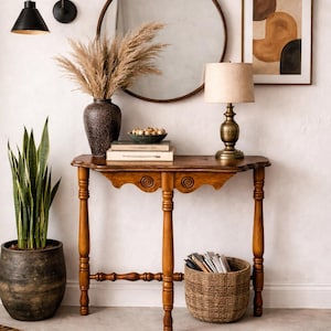May include: A warm-toned interior scene featuring a wooden console table with a lamp, books, and a woven basket. A large round mirror and artwork hang on the wall. A potted plant and decorative vase with dried grasses add to the decor.