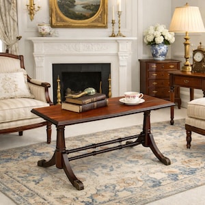 May include: A dark wood coffee table with a rectangular top and ornate legs. Two books and a magnifying glass rest on the table, along with a teacup and saucer. The table is set on a patterned rug in a living room setting.