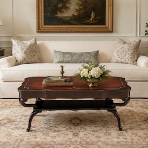 May include: A rectangular dark wood coffee table with curved legs and a lower shelf. The tabletop features a smooth, polished surface with a decorative inlaid border. A brass candlestick, two stacked books, and a bowl of white flowers sit on top.