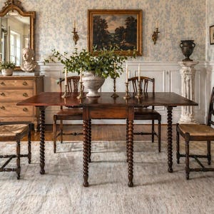 May include: A dark wood dining table with turned legs and a rectangular top. The table is surrounded by wooden chairs with woven seats. A large vase with greenery sits in the center, with candles and a painting on the wall.
