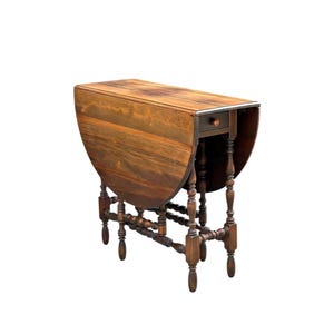 May include: A brown wooden drop-leaf table featuring a rectangular top with two hinged side leaves that fold downward. The table includes a single small drawer with a round knob and is supported by eight turned legs connected by stretchers.