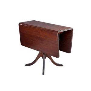 May include: A dark brown, antique drop-leaf table with a three-legged base. The table is made of wood and has a rectangular top with drop leaves that can be raised or lowered. The table is approximately 30 inches long when the leaves are down.