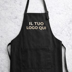 Puede incluir: Un delantal negro con una correa para el cuello con hebilla ajustable, un bolsillo frontal grande y el texto "IL TUO LOGO QUI" impreso en beige claro en el pecho, con el texto "Ricamo professionale Personalizzabile con if tuo logo" en la parte inferior.