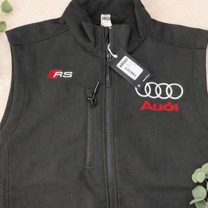 May include: Black vest with a zip-up front, featuring the Audi logo in white and red, and the letters "RS" in red. The vest has a small pocket on the chest and a tag attached. The vest is sleeveless.