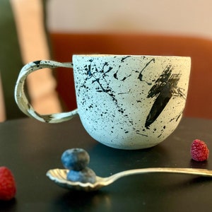 May include: A white ceramic mug with black splatter paint and a black brushstroke design. The mug is sitting on a black surface with a spoon and blueberries.
