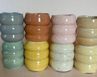 Puede incluir: Ocho tazas de cerámica dispuestas en dos pilas de cuatro. Las tazas presentan un diseño ondulado y acanalado con un acabado brillante. Los colores incluyen azul, verde, naranja y amarillo sólidos, junto con variantes moteadas en rosa, crema y bronceado.