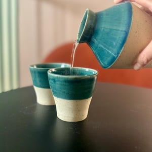 May include: Two teal and white ceramic mugs with a teal ceramic pitcher pouring liquid into one of the mugs. The mugs and pitcher have a speckled white glaze.