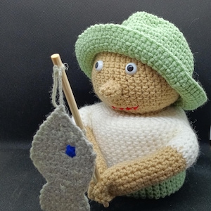 May include: A crocheted toy fisherman wearing a green hat and holding a wooden fishing rod with a gray crocheted fish attached. The toy has brown yarn for skin and white yarn for a shirt.