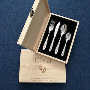 May include: A wooden box with a set of silver cutlery, including a fork, knife, and two spoons. The box is open, revealing the utensils against a black velvet lining. The box lid has the name "Elina" engraved, along with a date and text.