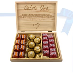 Op de afbeelding: Een houten doos met een hartvormig ontwerp op de deksel. De deksel is open en de doos is gevuld met verschillende chocolaatjes. De chocolaatjes zijn in twee rijen gerangschikt, waarbij de bovenste rij een selectie chocolaatjes bevat en de onderste rij een selectie chocolaatjes bevat. De doos is voorzien van de tekst "Liebste Oma" en een bericht waarin de ontvanger gefeliciteerd wordt met haar 75e verjaardag.