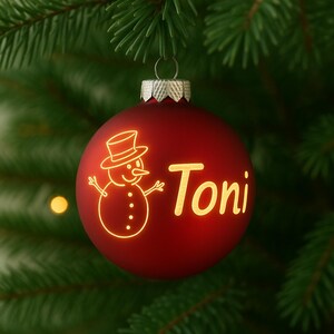 May include: A red Christmas bauble with the name "Toni" in gold lettering and a snowman illustration. The bauble hangs from a silver cap against a backdrop of green pine needles.