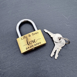 Könnte beinhalten: Ein goldfarbenes Vorhängeschloss mit silbernem Bügel und zwei Schlüsseln. Auf dem Vorhängeschloss stehen die Namen "Lilly & Sven", das Wort "love" und das Datum "24.03.2024". Die Schlüssel sind an einem silbernen Ring.