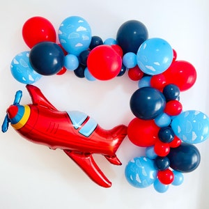 May include: A red airplane balloon with blue and yellow details, surrounded by a garland of blue, red, and black balloons. Some of the balloons have a white cloud design.
