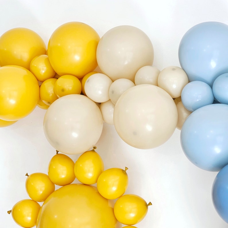 May include: A garland of balloons in yellow, white, and blue. The balloons are arranged in a circular shape, with the yellow balloons in the center and the white and blue balloons surrounding them.