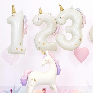 Könnte beinhalten: Weiße Ballons in Einhornform und Zahlenballons in Form der Zahlen 1, 2 und 3. Die Ballons haben goldene Hörner, rosa und lila Mähnen und goldene Sternakzente. Ein rosa herzförmiger Ballon ist ebenfalls vorhanden.