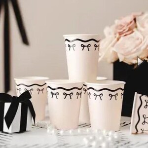 Puede incluir: Vasos de papel rosa pálido con un diseño de lazo negro alrededor del borde. Una caja de regalo en blanco y negro con un lazo está en primer plano. Un ramo de rosas rosas está en el fondo.