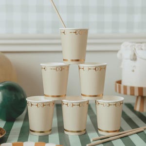 May include: A stack of six cream-colored paper cups with gold trim and a repeating gold equestrian bit design. One cup has a paper straw. The cups are arranged on a striped surface, suggesting a party or event setting.