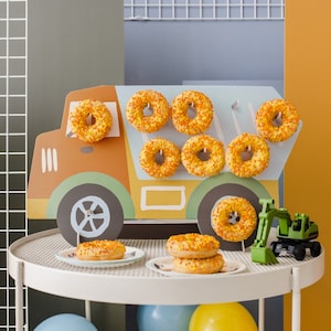 Puede incluir: Un camión de juguete de madera con forma de mezcladora de cemento con un esquema de pintura azul y amarillo. El camión está lleno de donuts con chispas naranjas. El camión está sobre una mesa blanca con una excavadora de juguete al lado.